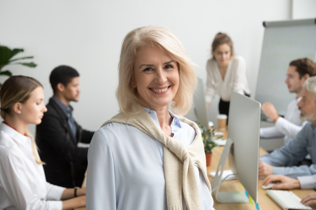 Smiling female aged company executive or team leader looking at camera, happy senior businesswoman teacher coach posing with office people at background, friendly older woman boss head shot portraitの写真素材
