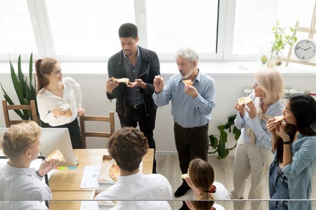 Multiracial young and senior colleagues eating pizza and talking at corporate lunch in coworking office, friendly diverse project team staff people enjoying meal at break discussing new ideas togetherの写真素材