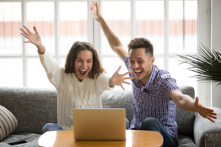 Young couple feeling excited by online win looking at laptop screen, happy man and woman celebrating victory watching video, screaming enjoying good news, motivated by achievement or great resultの写真素材