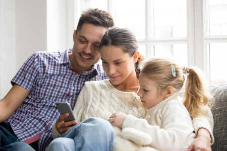 Parents with daughter watching video on mobile phone at home, young family couple with little girl...