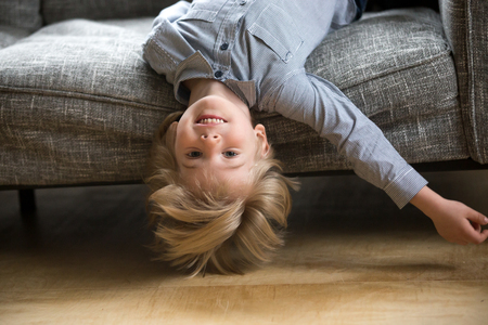 Cute funny boy lying upside down on sofa looking at camera, smiling playful preschool child having fun at home on couch, head shot portrait of happy blue-eyed kid with light hair playing at homeの写真素材