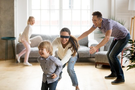 Blindfolded mother catching little son playing hide and seek at home, happy children having fun with...