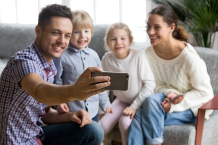 Smiling father holding smartphone taking selfie portrait of happy family with kids bonding together, husband making photo or shooting video of wife and children on mobile front camera, focus on phoneの写真素材