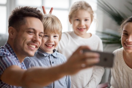 Kid sister making little cute brother bunny ears while father taking happy family selfie, funny children boy and girl having fun posing for self-portrait with smiling mom and dad bonding togetherの写真素材