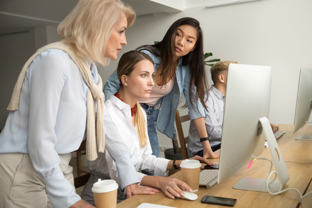 Serious multiracial female colleagues working together on online project, young and senior women employees helping in office teamwork, asian caucasian businesswomen collaborating focused on computerの写真素材