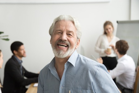Happy senior businessman laughing looking at camera in office, cheerful team leader posing with employees at background, aged teacher, old gray-haired boss or professional mentor head shot portraitの写真素材