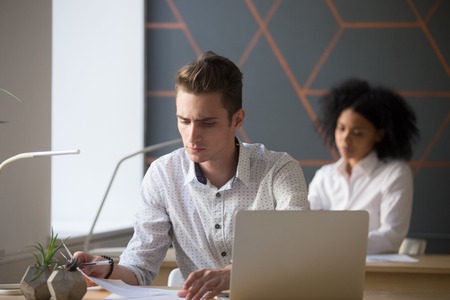 Serious millennial businessman attentively reading document focused on thinking in multiracial office, concerned employee analyzing report or contract, student preparing for test checking exam paperの写真素材