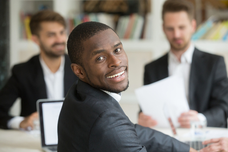 Smiling successful african american looking at camera back over shoulder while sitting at business meeting. Workers discussing company plans and strategies at briefing in background. Diverse team coworkingの写真素材