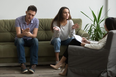 Stressed wife discussing relationship problems with female psychologist, explaining thoughts, husband indifferent, couch listening attentively. Couple getting help to save marriage, avoid break upの写真素材