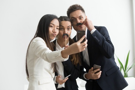 Coworkers trying mustache accessory, making silly faces posing to camera while making selfie on smartphone. Colleagues having fun at work, playing childish and taking pictures during work break.の写真素材