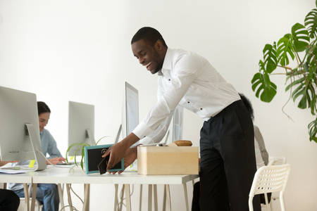 Happy african new employee unpacking box with belongings at workplace on first working day concept, excited black hired worker newcomer smiling holding framed picture settling in office on deskの写真素材