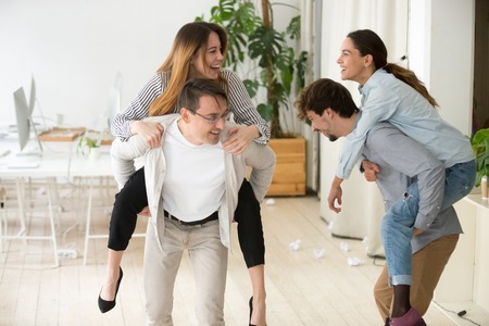 Happy colleagues having fun together enjoying piggyback ride in office, male employees holding women...