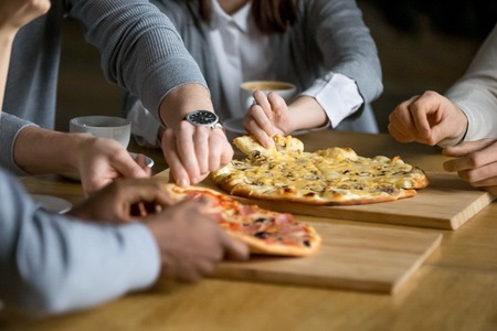 Hands of diverse people taking pizza slices from wooden board dining together, multiracial friends...