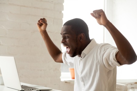 Excited amazed black worker throwing hands up, screaming celebrating online win, earning money, reaching goal, getting loan. Happy African American feeling euphoric after company business breakthroughの写真素材
