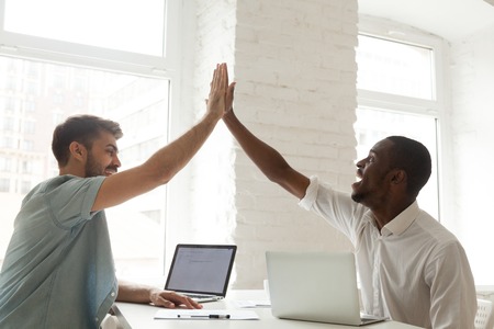 Excited African American worker giving high five to happy Caucasian colleague celebrating closing...