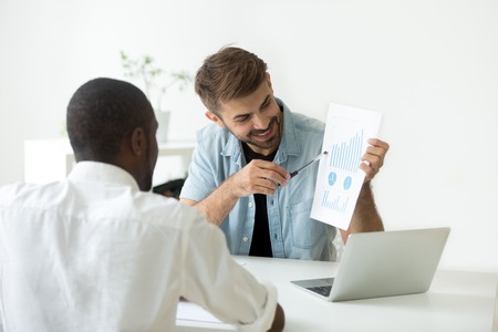 Smiling Caucasian worker showing visual material to African American colleague, explaining diagrams, talking about investing, funds, growing sales stats, demonstrating successful business strategyの写真素材