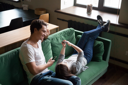 Smiling millennial couple talking holding smartphones relaxing on couch together, boyfriend and girlfriend checking social network news online or using mobile apps having fun with smartphones at homeの写真素材