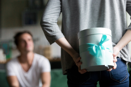 Woman hiding present behind back making romantic surprise for boyfriend or husband waiting for congratulation, rear close up view of female hands holding gift box, celebrating happy birthday conceptの写真素材