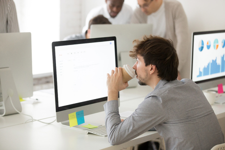 Thoughtful male professional drinking coffee, busy working at desktop computer, answering business emailの写真素材