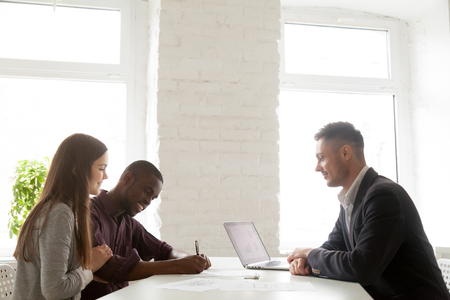 Multiethnic young spouses signing contract on property purchase while visiting real estate agent, African American husband putting signature on mortgage agreement. Concept of ownership, diversityの写真素材