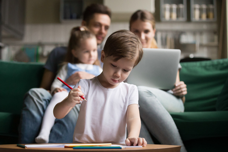 Preschool kid boy son drawing with colored pencils while parents with little sister using laptop on sofa in living room, family spending time together at home, creative child activities conceptの写真素材