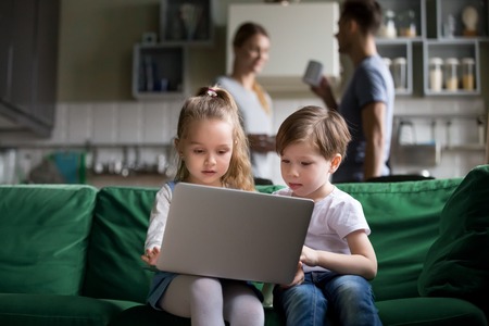 Cute children sister and brother watching online cartoons on laptop while parents talking in the kitchen, curious siblings boy girl using pc at home, kids computer addiction parental control conceptの写真素材