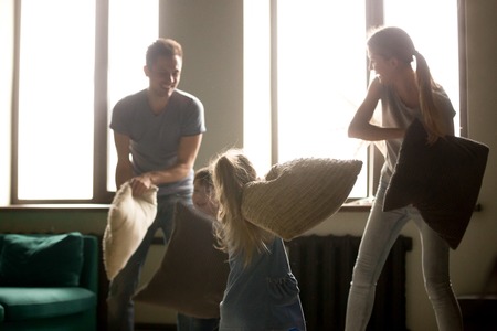 Happy parents and kids laughing having fun pillow fight indoors, excited smiling family playing together fighting cushions with little active children in living room, leisure activity at homeの写真素材