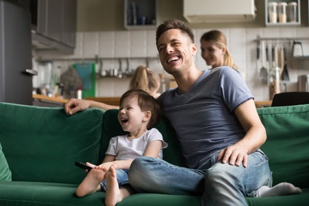 Happy dad and kid son holding remote control laughing at funny humor comedy film or tv show sitting on sofa at home, smiling father having fun with child boy watching television together on weekendの写真素材
