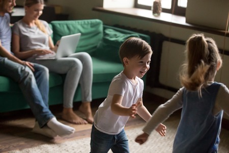 Happy siblings kids running playing funny game in living room while parents relaxing on sofa with laptop, little active boy brother and girl sister laughing having fun together at home on weekendの写真素材