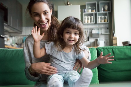 Portrait of happy excited young mother and kid girl waving hands looking at camera, smiling mom with child daughter making video call, family vloggers recording video blog or vlog together conceptの写真素材