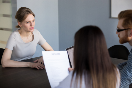 Serious millennial female job candidate talking during work interview in office, recruiters listening to applicant while hiring, hr team thinking considering employee candidature. Employment conceptの写真素材