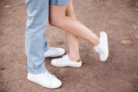 Close up of couple wearing jeans and white sneakers standing hugging and kissing outdoors in park, romantic young girlfriend playfully lift leg up reaching boyfriend embracing. Relationship conceptの写真素材