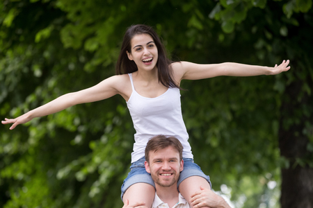Excited young girlfriend sitting on boyfriend shoulders stretching out arms pretending like flying during walk in park, happy millennial couple having fun outdoors, strong man carry playful womanの写真素材