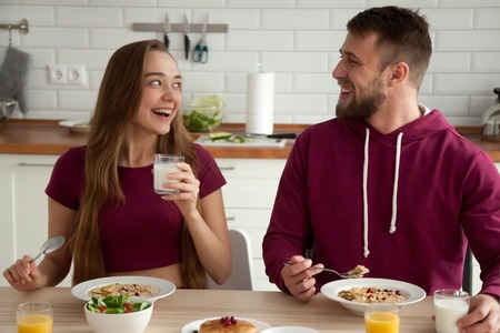 Happy young couple smiling enjoying delicious healthy oatmeal breakfast in kitchen, excited...