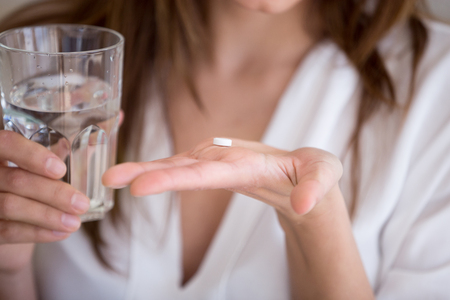 Woman holding pill and glass of water in hands taking emergency medicine, supplements or antibiotic antidepressant painkiller medication to relieve pain, meds side effects concept, close up viewの写真素材