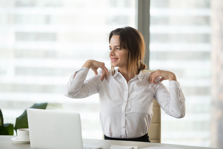 Smiling businesswoman doing easy office exercises to relieve neck and shoulder muscle tension from sedentary computer work, young employee taking break stretching for back relaxation at workplaceの写真素材