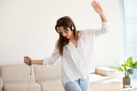 Carefree woman in headphones dancing to music playing on smartphone at home, young girl listening to new songs in mobile player app relaxing on weekend with new tracks podcast playing via phoneの写真素材