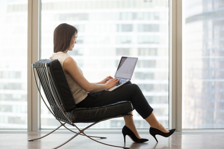 Young woman sitting in modern chair at home office using laptop for distance language learning studying online searching for internet business education course computer software, e-learning conceptの写真素材