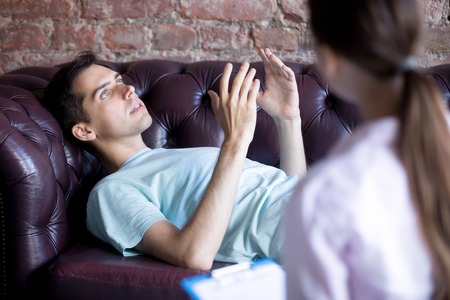 Young man at the psychotherapist, lying on couch, emotionally speaking, client discussing with female psychotherapist counselor his melancholy, mental health problemsの写真素材