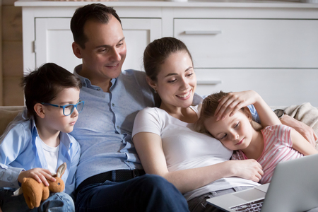 Happy young family of four sitting in living room spending time together, parents and kids relax watching cartoons on laptop, mom and dad cuddle with children hugging enjoying movie on computerの写真素材