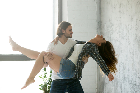 Strong young man holding happy girlfriend in hands carrying her to bed, excited millennial loving couple enjoying close intimate moments at home together, funny spouses hug dancing swirling in bedroomの写真素材