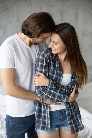 Loving affectionate man hug girlfriend from behind, romantic millennial couple embrace enjoying intimate moment together, boyfriend holding beloved girl in arms, standing touching, looking in the eyesの写真素材