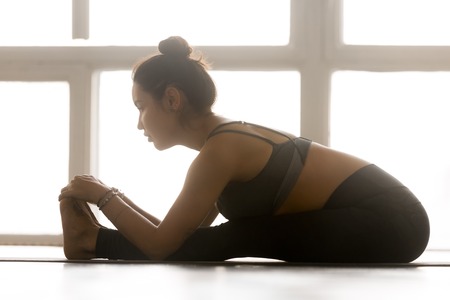 Young sporty attractive woman practicing yoga, doing Seated forward bend exercise, paschimottanasana pose, working out, wearing sportswear, grey pants, top, indoor full length, at yoga studioの写真素材