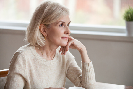 Thoughtful middle aged mature woman looking away drinking coffee tea thinking of future, dreamy beautiful senior lady relaxing alone dreaming hoping contemplating sitting at table lost in thoughtsの写真素材