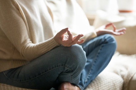 Calm senior woman sitting on sofa holding hands in mudra practicing home yoga in lotus pose for relaxation, old mature middle aged female enjoying meditating alone for peaceful mind, close up viewの写真素材