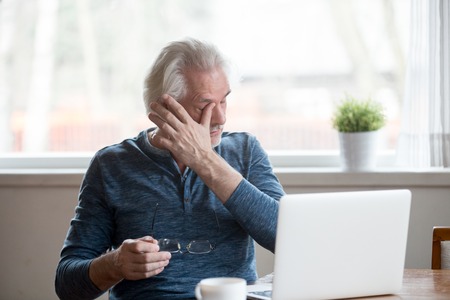 Fatigued mature old man taking off glasses suffering from tired dry irritated eyes after long computer use, senior middle aged male feels eye strain problem or blurry vision working on laptop at home