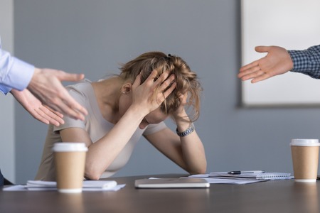 Stressed young businesswoman holding head in hands suffering from unfair gender discrimination at work concept, female employee frustrated by male colleagues bullying humiliating woman at workplaceの写真素材