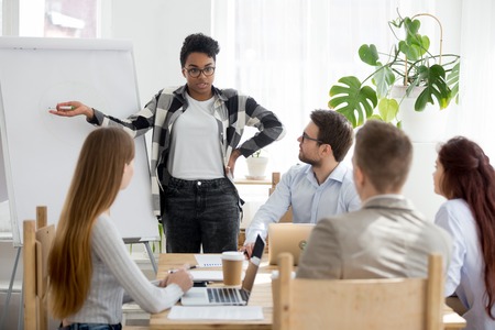 Businesspeople at seminar sitting in office. African leader boss female standing demonstrate show on whiteboard development strategy plan graph, argue with young colleagues clients at business meetingの写真素材