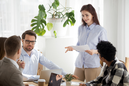 Diverse millennial businesspeople sitting at seminar in office boardroom. Coach leader woman or employee female hold report telling about sales statistics analysis to colleagues at business meetingの写真素材