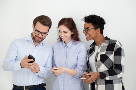 Group of people standing together opposite white wall looking on smartphone. Man showing to...